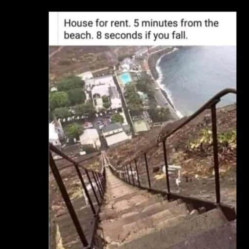 Architecture Memes, Dark Humor, Expectation vs Reality, Funniest Memes, Funny Sign Memes, Funny Text Memes, Realtor Memes, Rent Memes, Travel Memes, Vacation Memes, WTF Memes, The meme shows a dramatic, vertigo-inducing view from the top of a steep outdoor staircase plunging down a rocky hillside toward a coastal neighborhood and the ocean below. Metal railings frame the stairs, which appear narrow, uneven, and extremely long, making the descent look more like a survival challenge than a casual walk to the beach. At the top, the text reads: “House for rent. 5 minutes from the beach. 8 seconds if you fall.” The calm real-estate phrasing clashes with the alarming visual of a staircase so steep that one wrong step could turn the advertised beach access into a high-speed tumble.