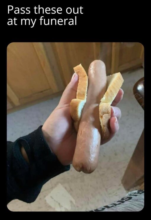 Dark Humor, Death Memes, Dirty Mind Memes, Double Meaning Memes, Food Memes, Funeral Memes, Funniest Memes, Innuendo Memes, NSFW, Penis Memes, WTF Memes, A black-background meme shows large white text reading, “Pass these out at my funeral,” above a close-up photo of someone holding a hot dog in a split bun. The hot dog is positioned upright and centered, with the bun pulled open on both sides like it is being presented dramatically.