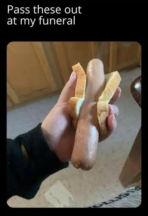 Dark Humor, Death Memes, Dirty Mind Memes, Double Meaning Memes, Food Memes, Funeral Memes, Funniest Memes, Innuendo Memes, NSFW, Penis Memes, WTF Memes, A black-background meme shows large white text reading, “Pass these out at my funeral,” above a close-up photo of someone holding a hot dog in a split bun. The hot dog is positioned upright and centered, with the bun pulled open on both sides like it is being presented dramatically.