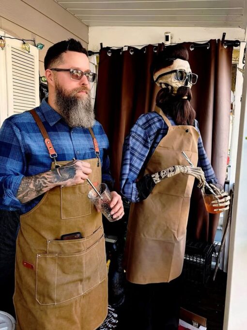 Beard Memes, Death Memes, Funniest Memes, Guy Memes, Halloween Memes, Spooky Memes, Weird Memes, A bearded man in glasses, a blue plaid shirt, and a tan work apron stands indoors holding a drink with a straw, looking calm and serious. Next to him is a life-size skeleton dressed almost exactly the same way: plaid shirt, tan apron, sunglasses, styled dark hair, and an enormous fake beard, also posed with a drink in hand.