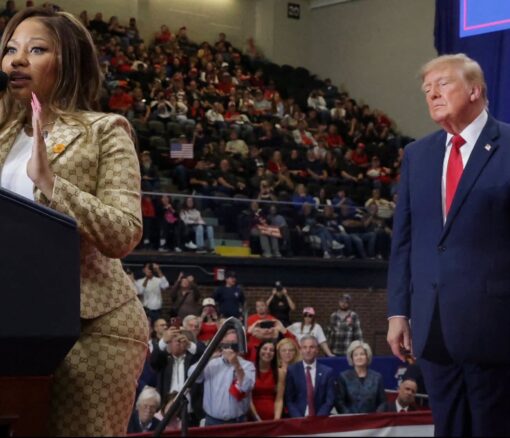 Celebrity Memes, Current Event Memes, Donald Trump Memes, Funniest Memes, MAGA Memes, Music Memes, News Memes, Political Memes, Pop Culture Memes, Rap Memes, Trump and Nicki’s Ass, The image shows a rally-stage scene inside a packed arena: Nicki Minaj stands at a podium on the left in a fitted, patterned gold outfit, while Donald Trump stands several feet behind her on the right in a dark suit and red tie. The background is filled with seated spectators, phones raised, flags visible, and a small black sign near the top that reads “30.”