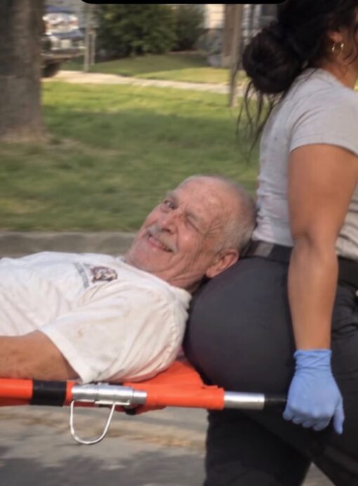 Awkward Memes, Butt Memes, Caught You Looking Memes, Dirty Mind Memes, Funniest Memes, Health Memes, Innuendo Memes, NSFW, Old People Memes, Pics Gone Wrong, WTF Memes, A candid-looking outdoor scene shows an elderly man lying on an orange stretcher while being carried or moved by a medical worker. His head is pressed right up against the backside of a woman in dark leggings, and he looks directly at the camera with a sly wink and grin, as if he knows exactly how ridiculous the moment appears. The only text on the image reads “NPLASMANENT,” an odd, jumbled-looking word that adds to the chaotic, accidental feel of the image rather than clearly explaining it.