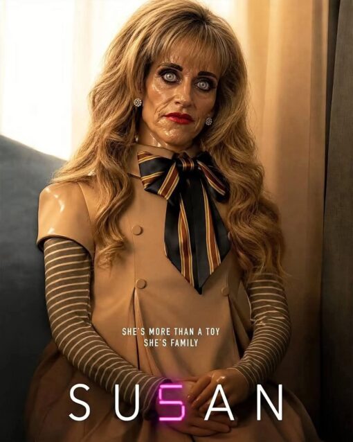 AI Memes, Creepy Memes, Family Memes, Funniest Memes, Horror Memes, Movie Memes, Pop Culture Memes, Robot Memes, Scary, Toy Memes, Weird Memes, The image is styled like a dramatic horror-movie poster: a lifelike blonde doll-woman stands stiffly in warm, dim lighting, staring ahead with glassy, unsettling eyes, heavy makeup, and a frozen expression. She wears a beige dress with striped sleeves and a large striped bow, giving her the polished but uncanny look of a toy that is almost human but not quite. Across her torso, small white text reads “SHE'S MORE THAN A TOY / SHE'S FAMILY,” while the bottom spells out “SUSAN” in large spaced-out letters, with the center letter glowing pink in a way that mimics the stylized title treatment of a sci-fi horror poster.